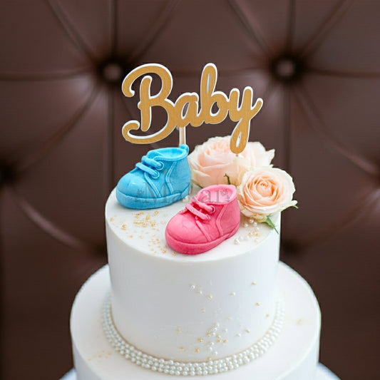 Elegant white cake decorated with edible blue and pink fondant baby shoe toppers, golden 'Baby' sign, soft peach roses, and pearl accents – perfect for baby showers and gender reveal celebrations.