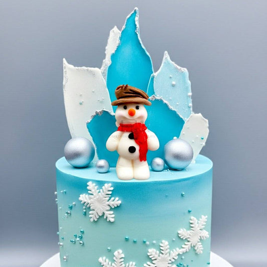 Blue winter cake decorated with a fondant snowman wearing a brown hat and red scarf, surrounded by snowflakes and silver baubles.