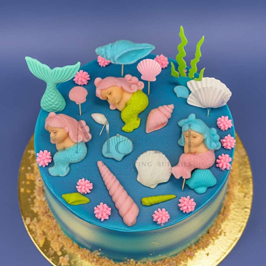 Blue ocean-themed cake decorated with pastel mermaid toppers, seashells, seaweed, and coral accents — part of the Mini Mermaid Cake Topper Set from Makai Cake Decorating Supplies.