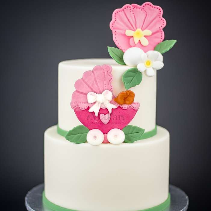 Two-tier white baby shower cake decorated with a pink fondant pram, green leaves, and sugar flowers, perfect for a girl’s celebration – Makai Cake Decorating Supplies.