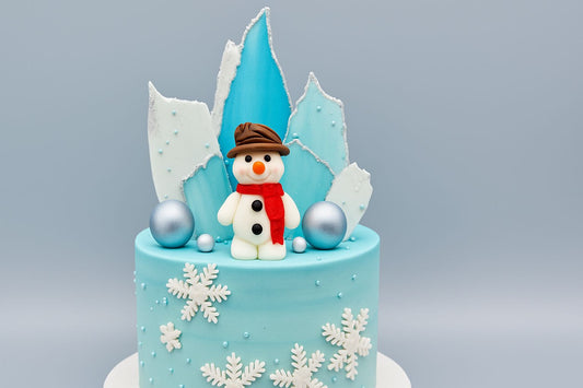 Winter-themed cake decorated with a fondant snowman, blue and white ice shard toppers, silver balls, and white snowflakes on a light blue background.