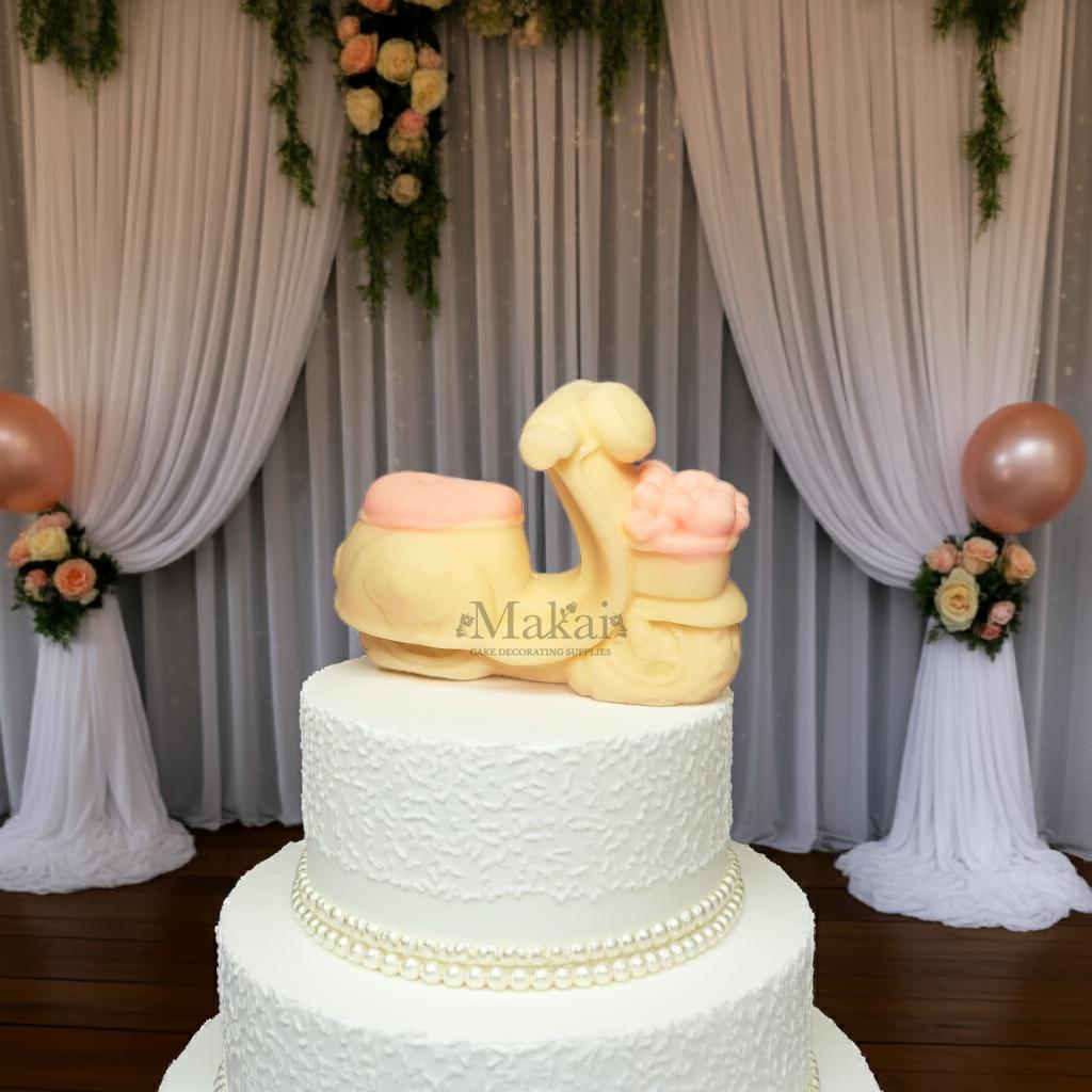 Edible scooter cake topper with pink flowers, displayed on a white wedding cake with elegant draped backdrop – Makai Cake Decorating Supplies