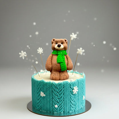 Winter cake with a fondant bear in a green scarf, surrounded by delicate snowflakes and a knitted-style blue icing design.