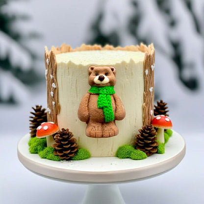 Winter forest-themed cake featuring a cute fondant bear wearing a green scarf, surrounded by pine cones and mushrooms on a snowy base.