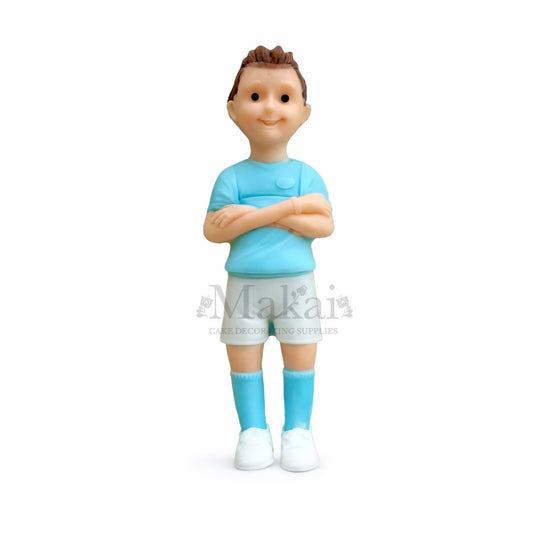 Figurine of a cake topper wearing a blue shirt and shorts with arms crossed on a white background.