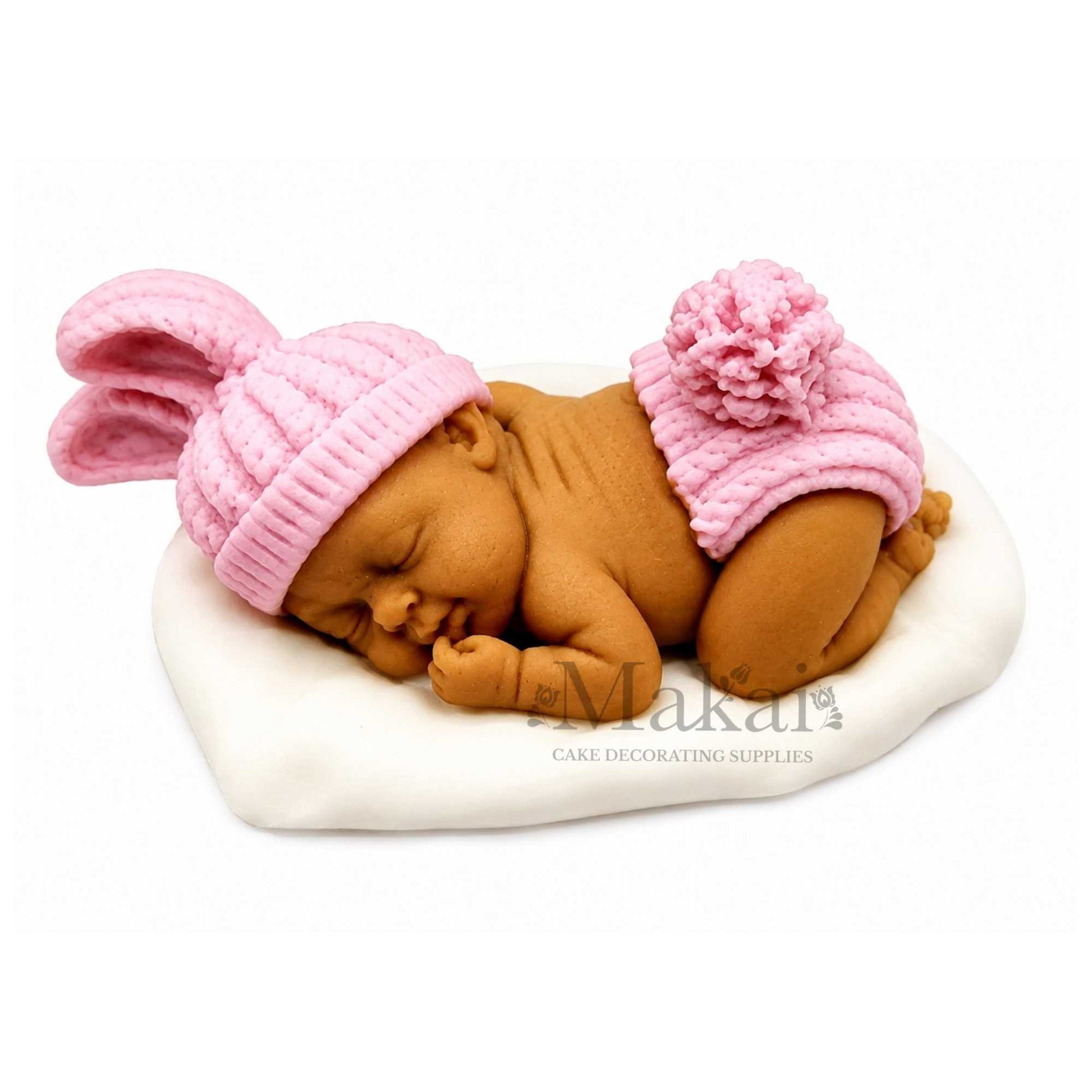 Fondant sleeping baby cake topper with darker skin tone, wearing a detailed pink bunny hat and matching knitted-style shorts.