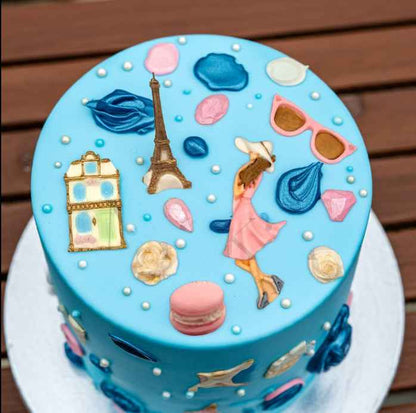 Paris themed blue birthday cake with edible fondant decorations including Eiffel Tower, woman in pink dress, macarons, sunglasses and flowers.