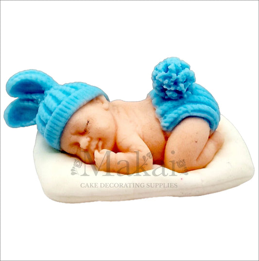 Sleeping baby boy cake topper with blue hat and diaper resting on a white cushion.
