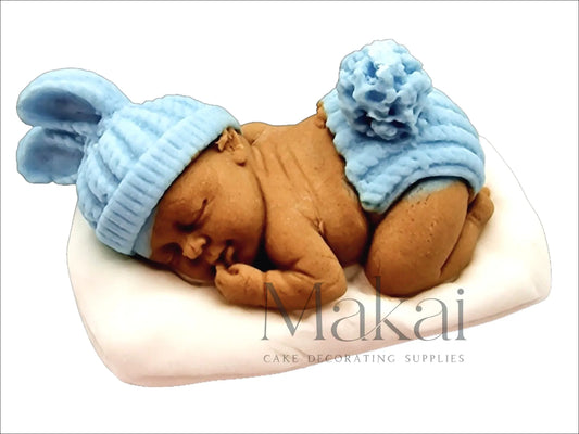 Darker-skinned fondant sleeping baby cake topper with a blue hat and little briefs, lying on a white pillow.