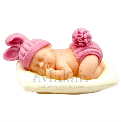 Sleeping baby girl cake topper with pink hat and diaper on a white cushion.