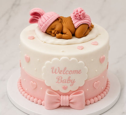 A round white and pink baby shower cake decorated with a fondant sleeping baby figure wearing a pink hat and diaper, with a pink bow and 'Welcome Baby' plaque on a marble surface.
