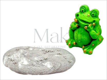 Edible smiling green frog cake topper with gold details sitting in front of a grey stone
