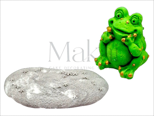 Edible smiling green frog cake topper with gold details sitting in front of a grey stone